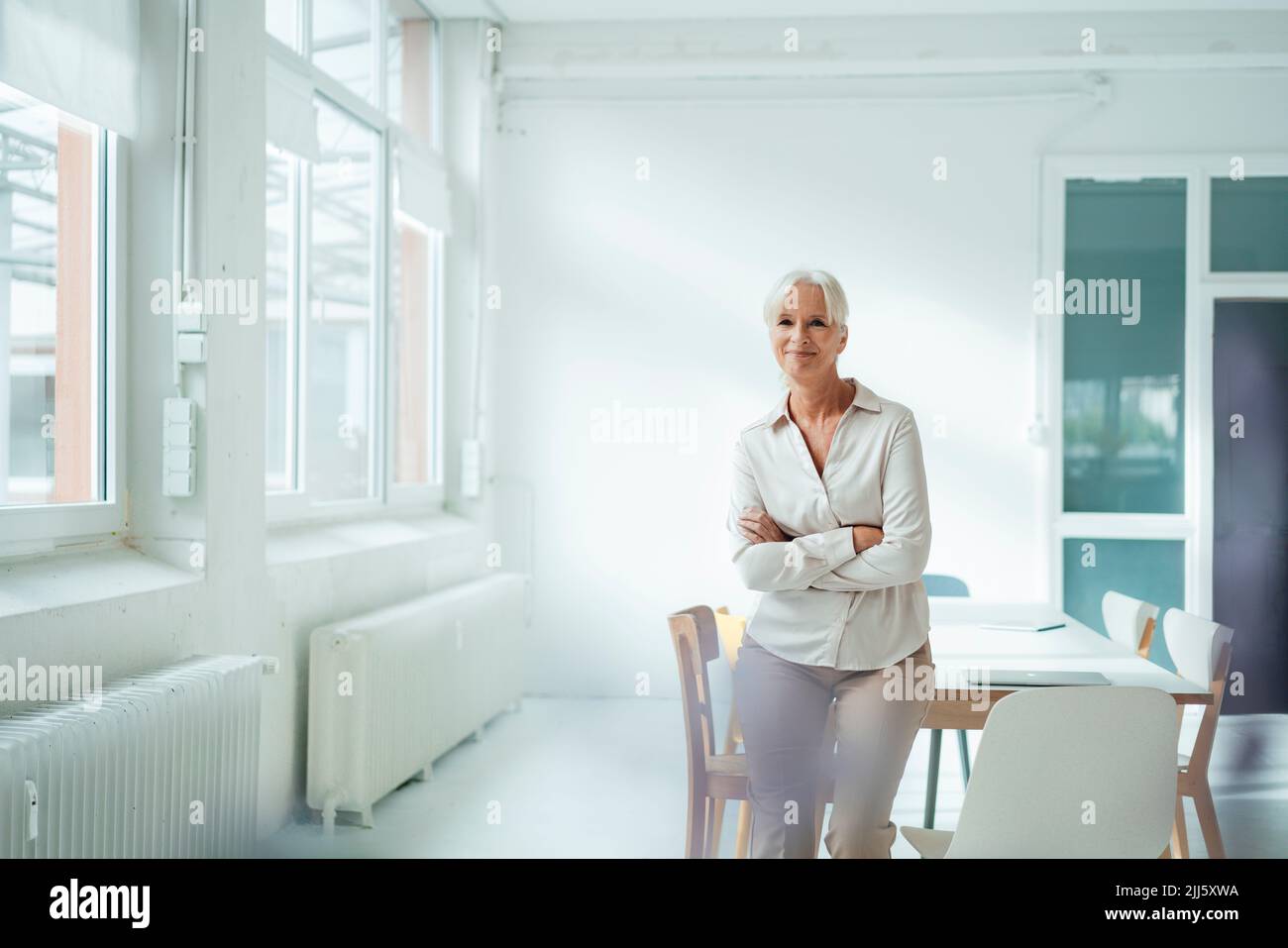 Smiling businesswoman with arms crossed leaning on desk in office Stock Photo