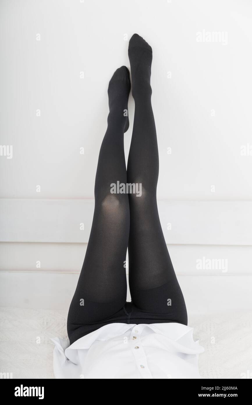 Beautiful long female legs in stockings. Girl putting on stockings at home in a white room. Black tights. Varicose veins prevention. Woman body in Stock Photo