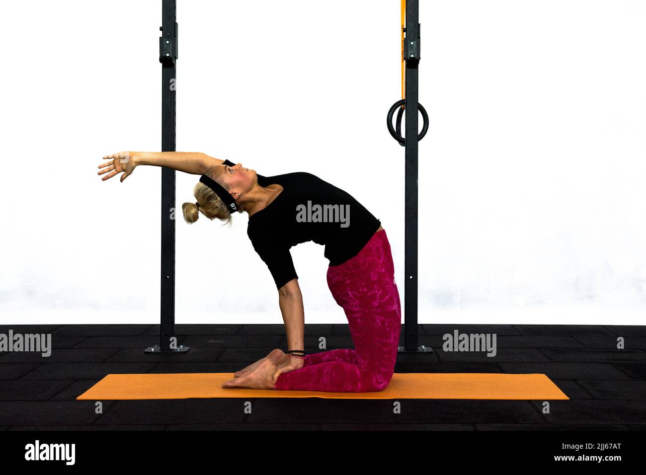 A youth lady trainer is performing the Camel yoga pose in a Gym with one arm upwards. Ustrasana the camel pose for lower back and throat stimulation. Stock Photo