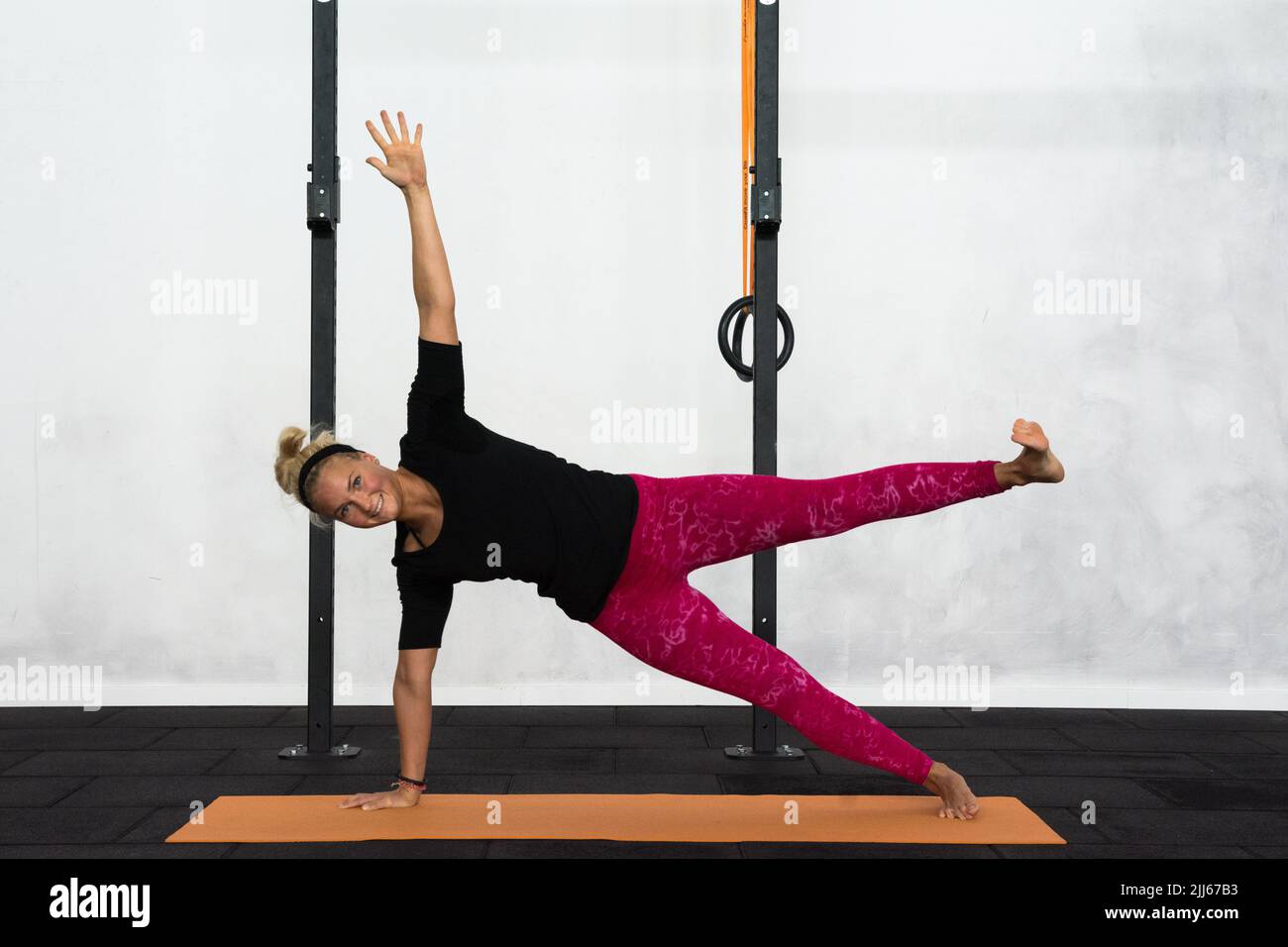 A youth blonde lady performing side plank with one leg up yoga pose. Vashistasana with one leg up yoga posture for balance, arms and legs strengthen. Stock Photo