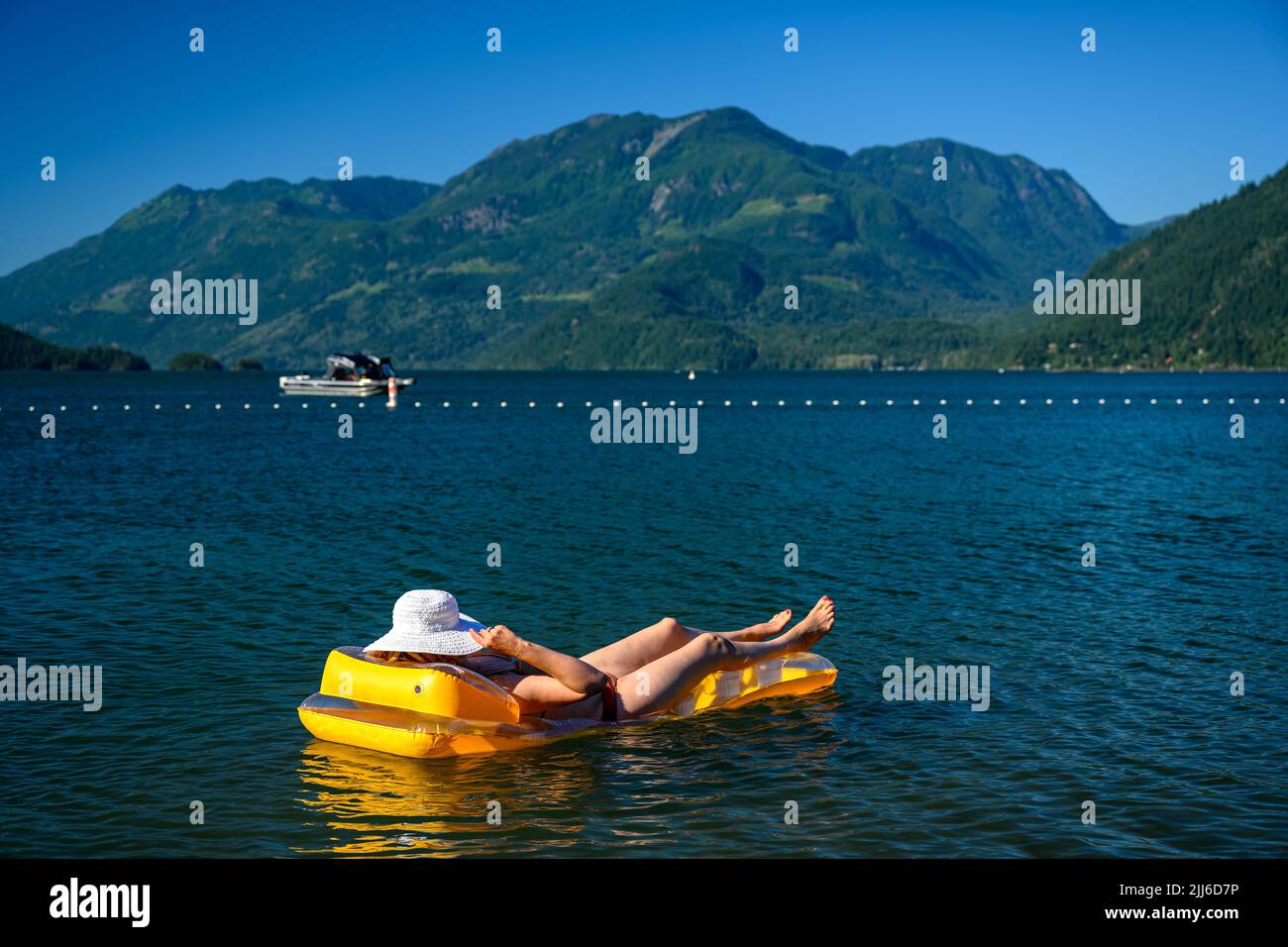 Mid adult woman lying on inflatable mattress, wearing a white straw sun hat that obscure her face while floating on the beautiful Harrison Lake surrou Stock Photo