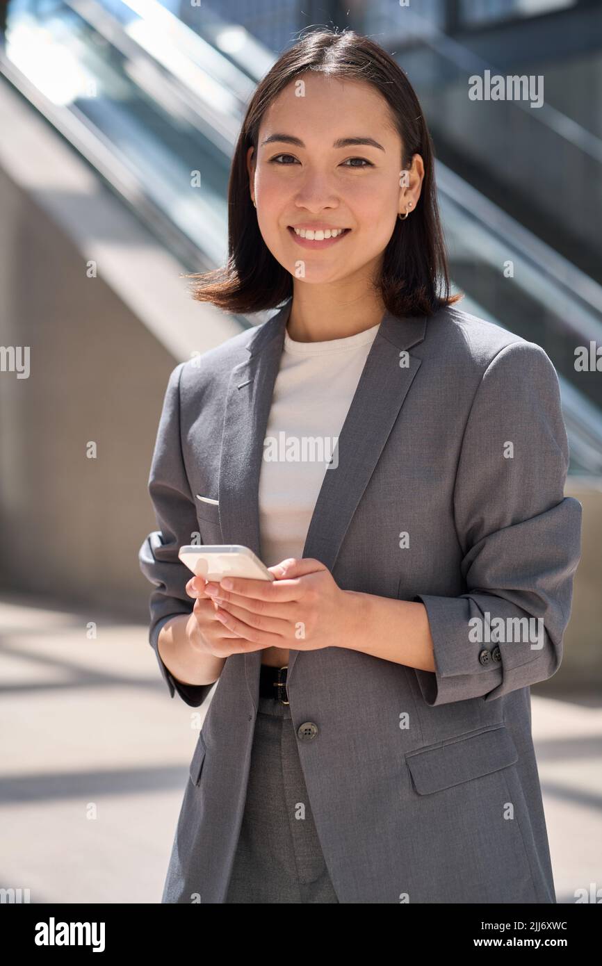 Smiling young Asian businesswoman standing on urban city street using ...