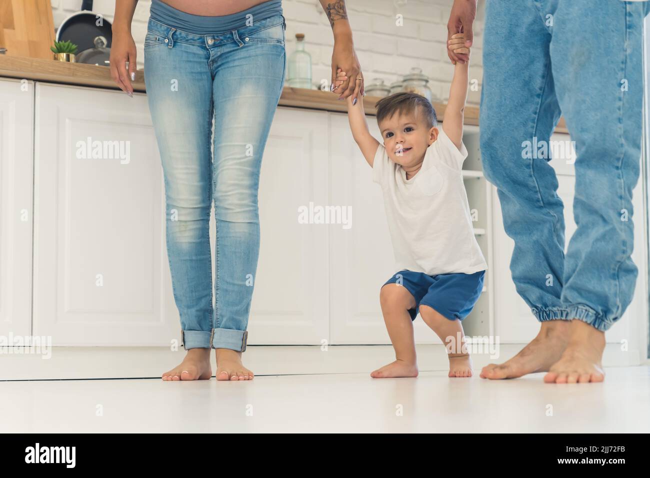 Family of three walking barefoot in their kitchen. Toddler boy standing between his parents and holding their hands in order to jump up. High quality photo Stock Photo