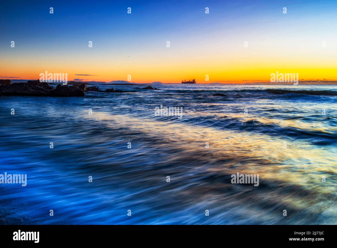 Blurred waves on Pebbly beach in Forster town on Pacific coast of Australia with distant ship on horizon at sunrise. Stock Photo