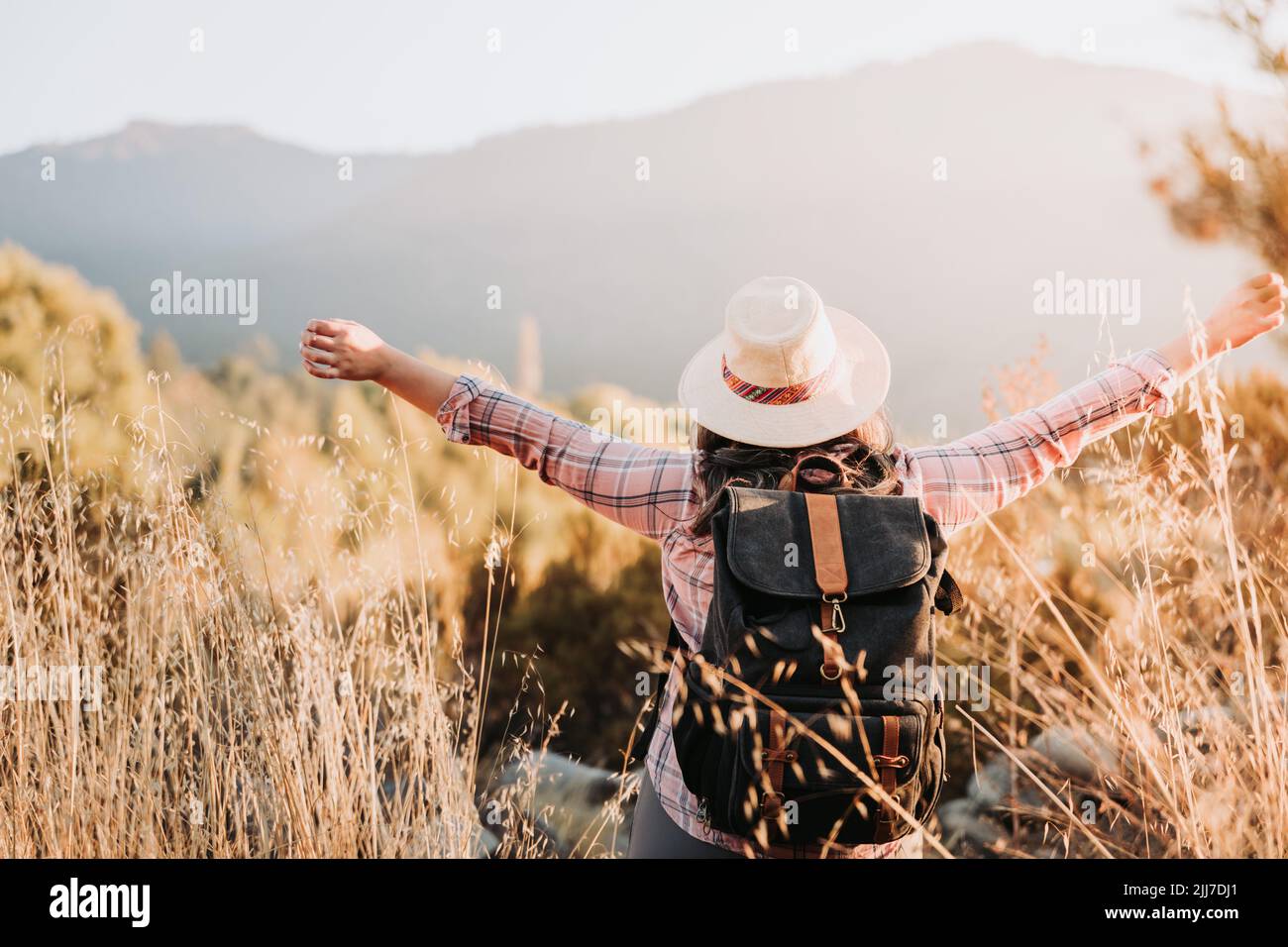 Unrecognizable woman with a hat and a backpack on, raising her arms in victory in nature. Stock Photo