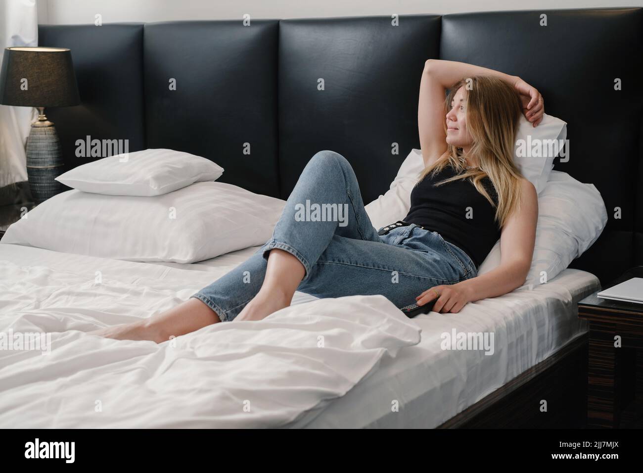 woman in hotel room bed. Relaxed young Female having rest in modern hotel while traveling. Watching TV with remote control in the morning. Copy space Stock Photo