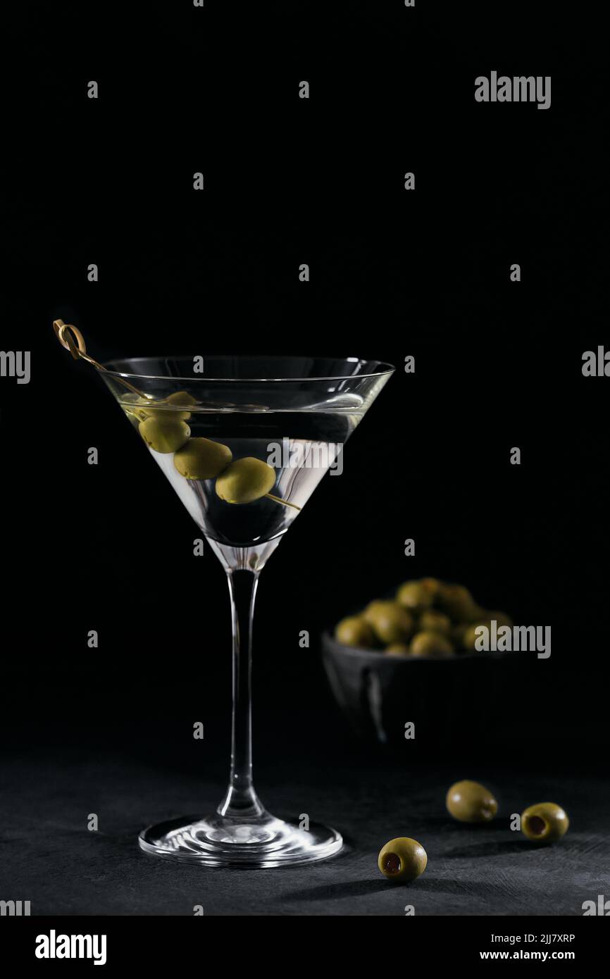 Glass of classic dry martini cocktail with olives on dark stone table against a black background