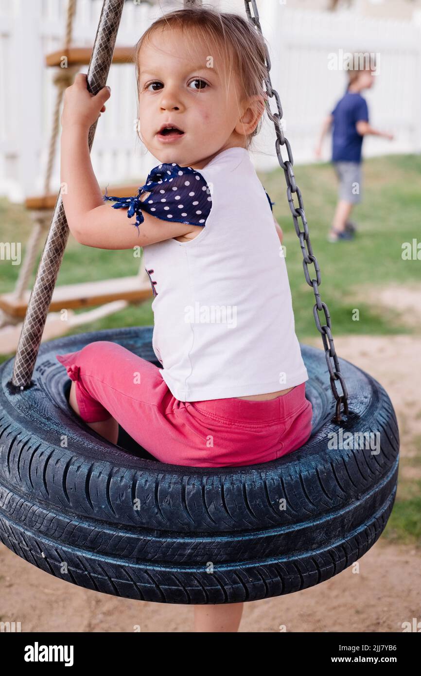 Child swinging on the rings, swings from wheels on playground. Girl having fun during walk. Strong, sporty and feet in summer Stock Photo