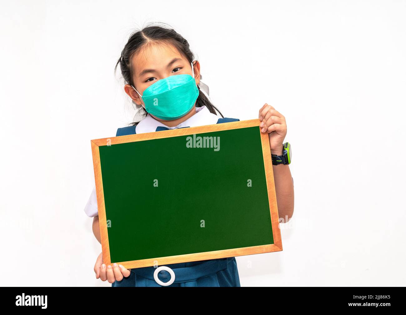 Portrait Asian student child in identified school uniform, holding small blackboard or chalkboard, wearing face mask, isolated image on white backgrou Stock Photo