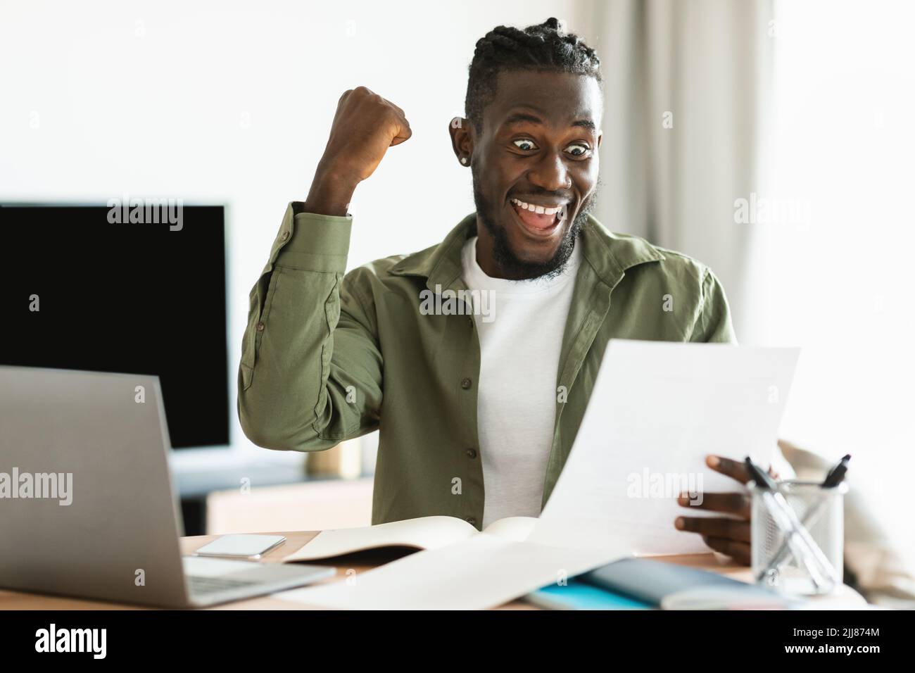 Good news. Overjoyed black man reading letter, raising fist and exclaiming with excitement, sitting at home office Stock Photo