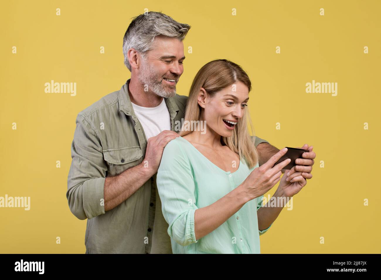 Excited middle aged spouses looking at cellphone screen, using new application, yellow studio background Stock Photo
