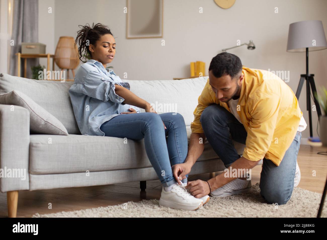 Black Husband Caring For Pregnant Wife Lacing Her Shoes Indoor Stock Photo