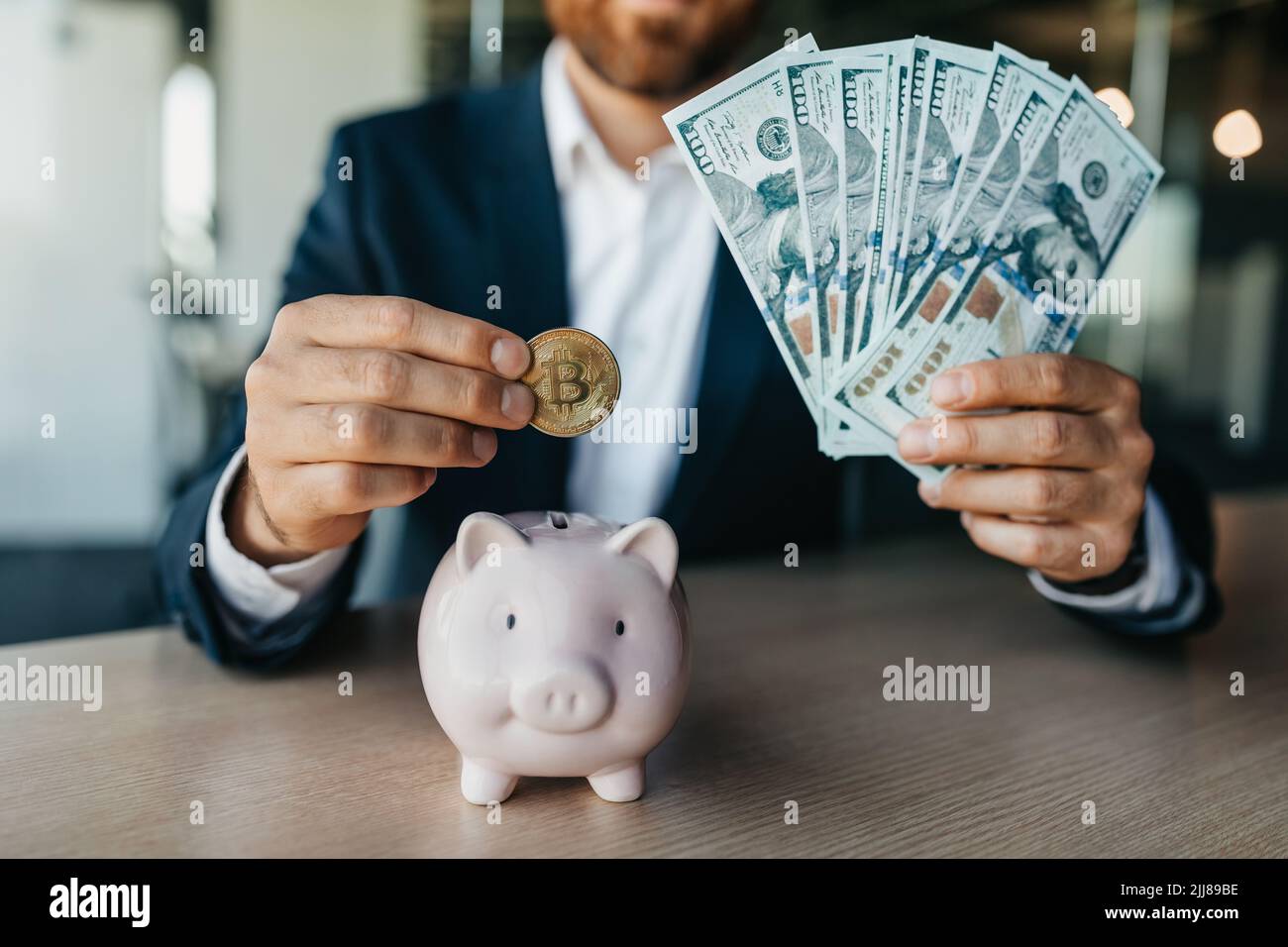 Successful businessman putting bitcoin coin in piggybank and holding money cash, sitting at table in office Stock Photo