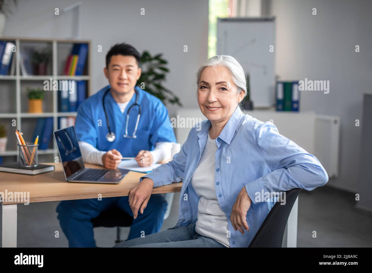 Smiling elderly european woman patient on meeting with young asian doctor in uniform look at camera Stock Photo