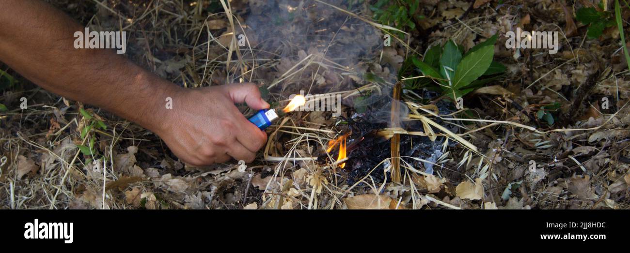 Image of the hand of an arsonist who starts a fire in a wood with a lighter. Reference to the problem of forest fires. Horizontal banner Stock Photo