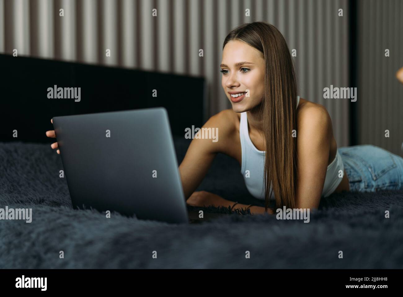 Smiling woman catching up on her social media as she relaxes in bed with a laptop computer on a lazy day Stock Photo