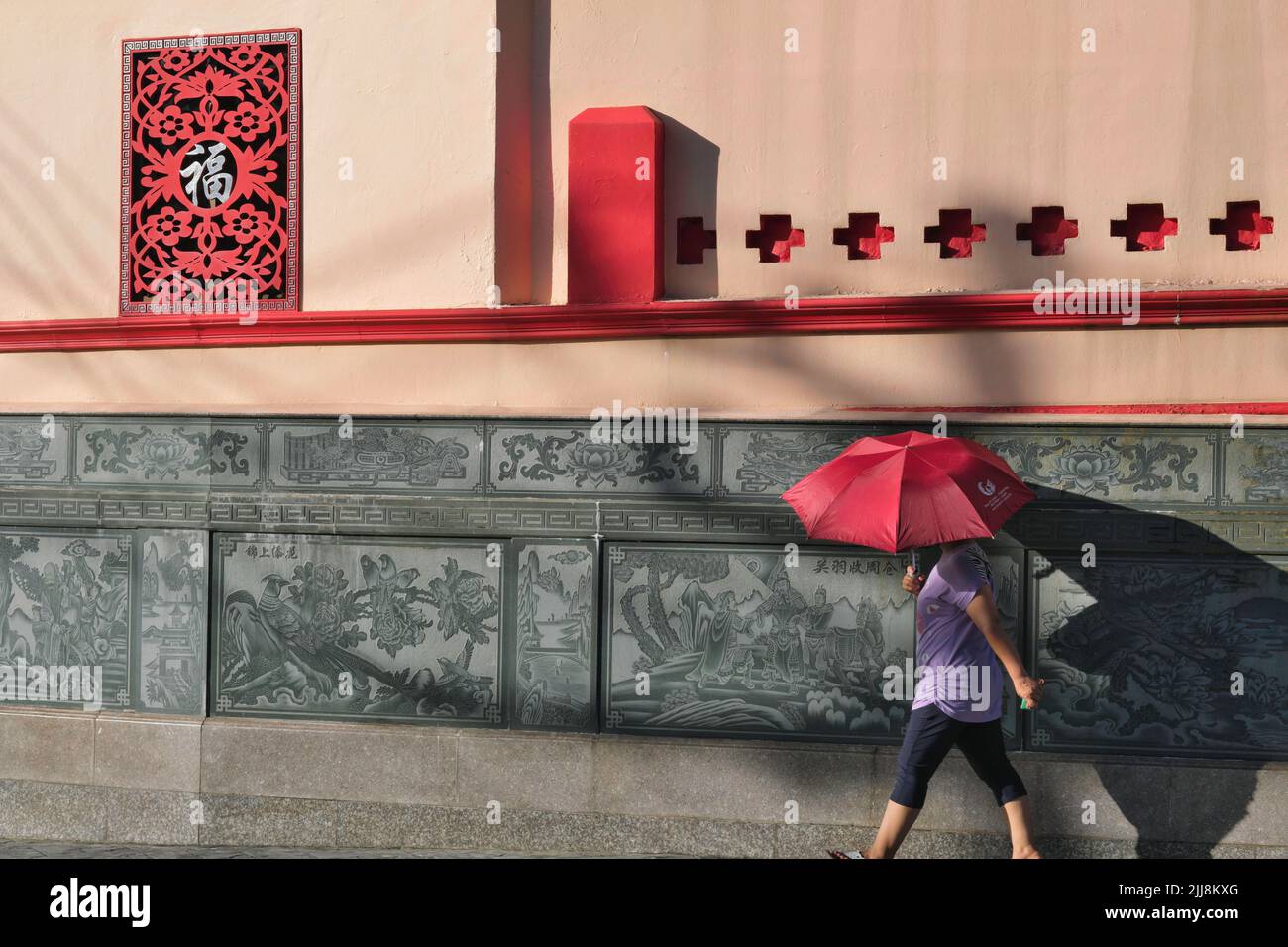 A woman with a red umbrella passes a Chinese-style frieze outside (Taoist) Pud Cho Shrine in Phuket Town, Thailand, casting a shadow on the wall Stock Photo