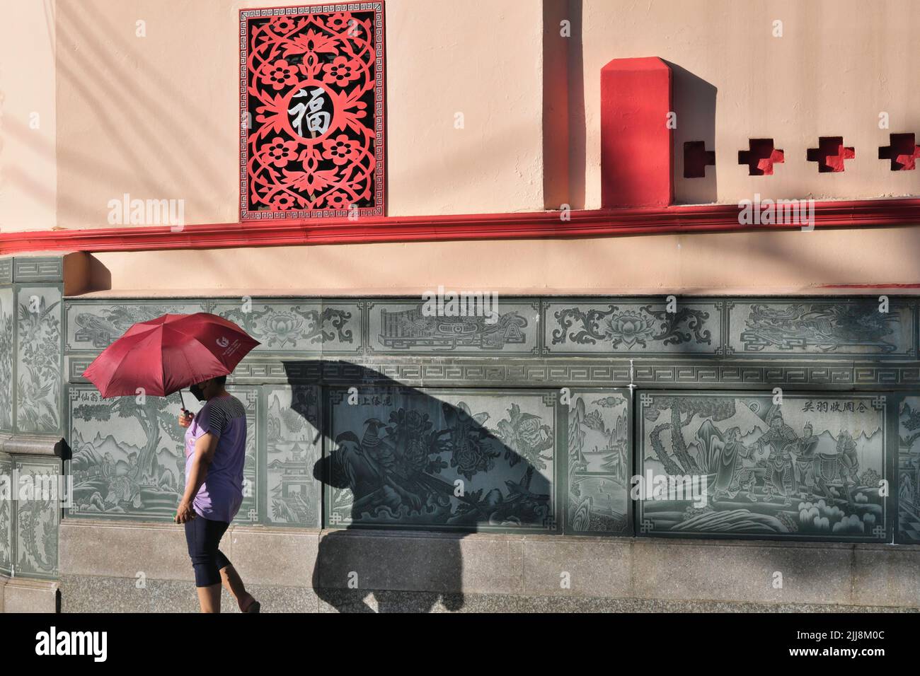 A woman with a red umbrella passes a Chinese-style frieze outside (Taoist) Pud Cho Shrine in Phuket Town, Thailand, casting a shadow on the wall Stock Photo