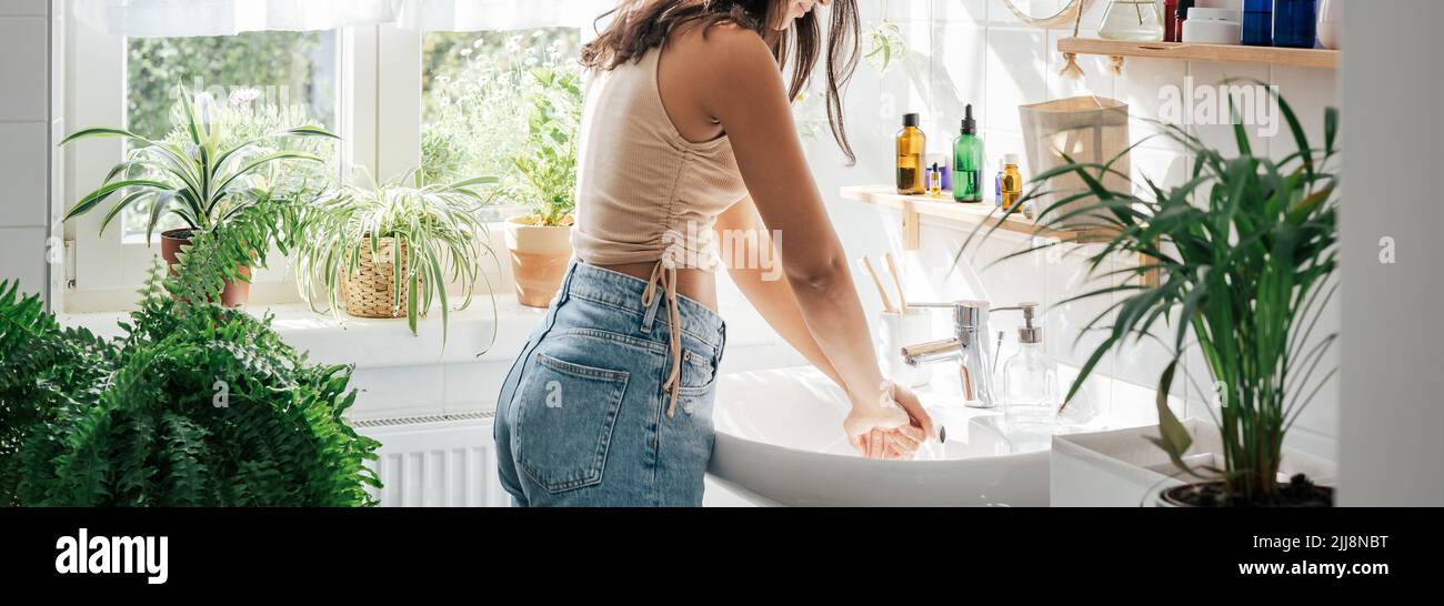 Young hispanic woman washing hands in white bathroom with biophilic design. Wellness concept. Self care. Long banner Stock Photo