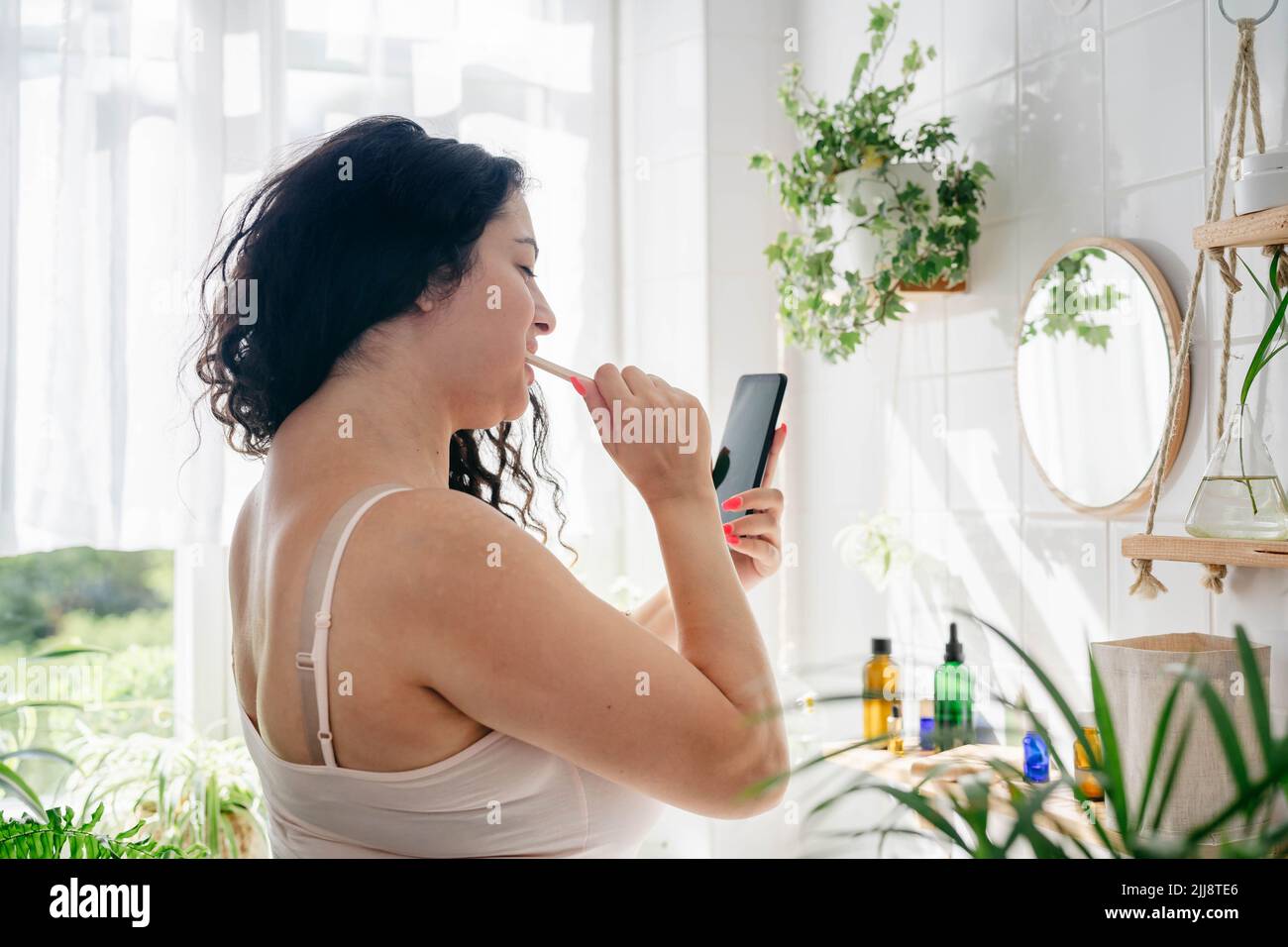 Young beautiful woman with overweight brushing her teeth and looking at the phone in light batroom with many green plants. Wellness, everyday technology Stock Photo
