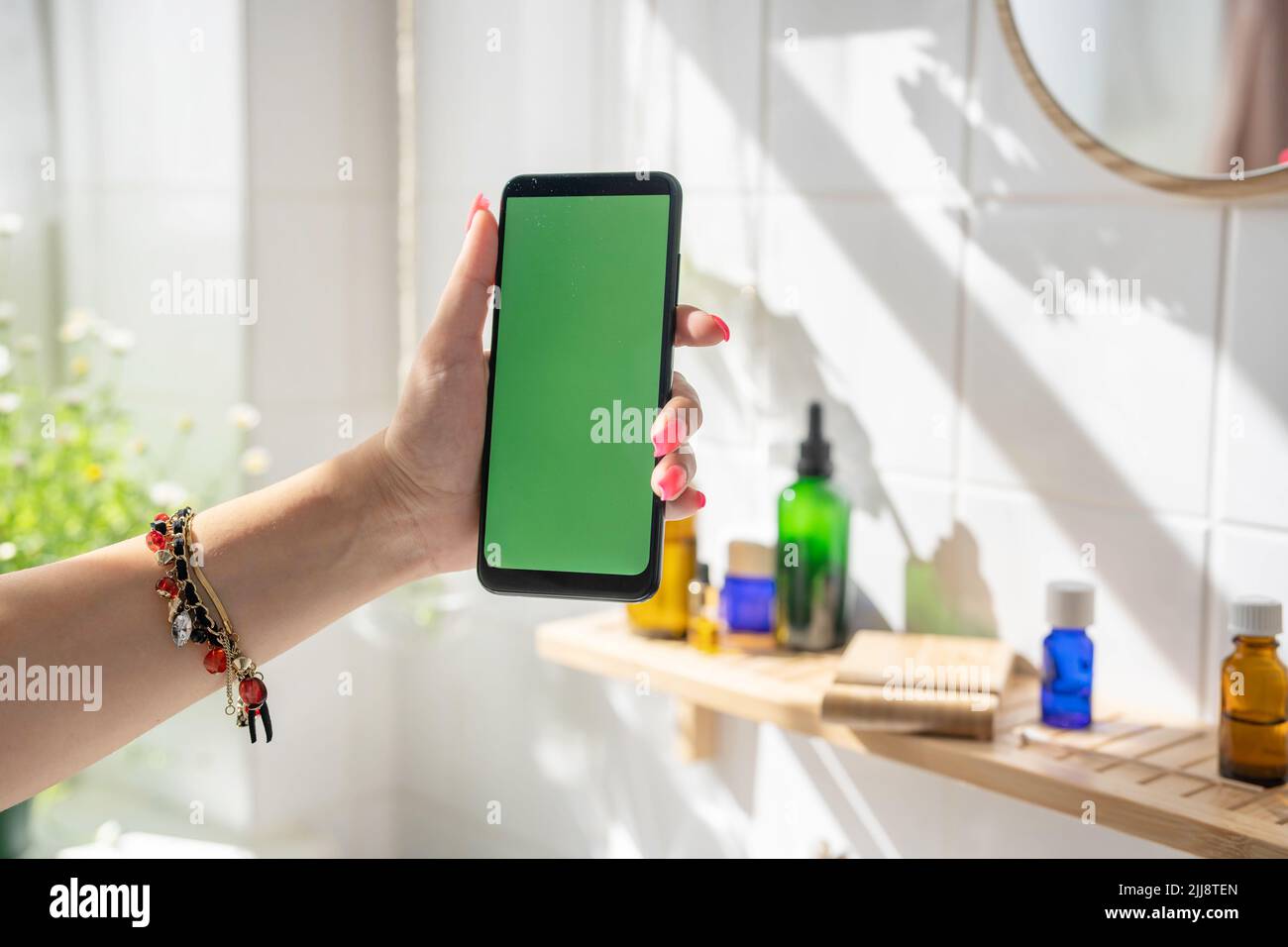 Female hand holding smartphone with green screen in bathroom against bamboo shelf with glass cosmetic bottles. Shadows from the sun on the white tiled wall Stock Photo