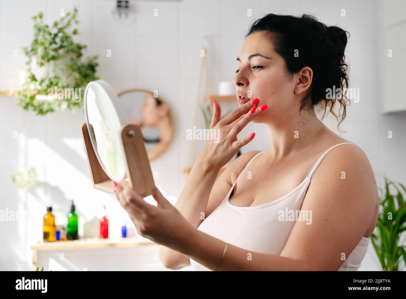 Young woman looking at the mirrow and examine her face in white bathroom. Wellness and body positivity Stock Photo