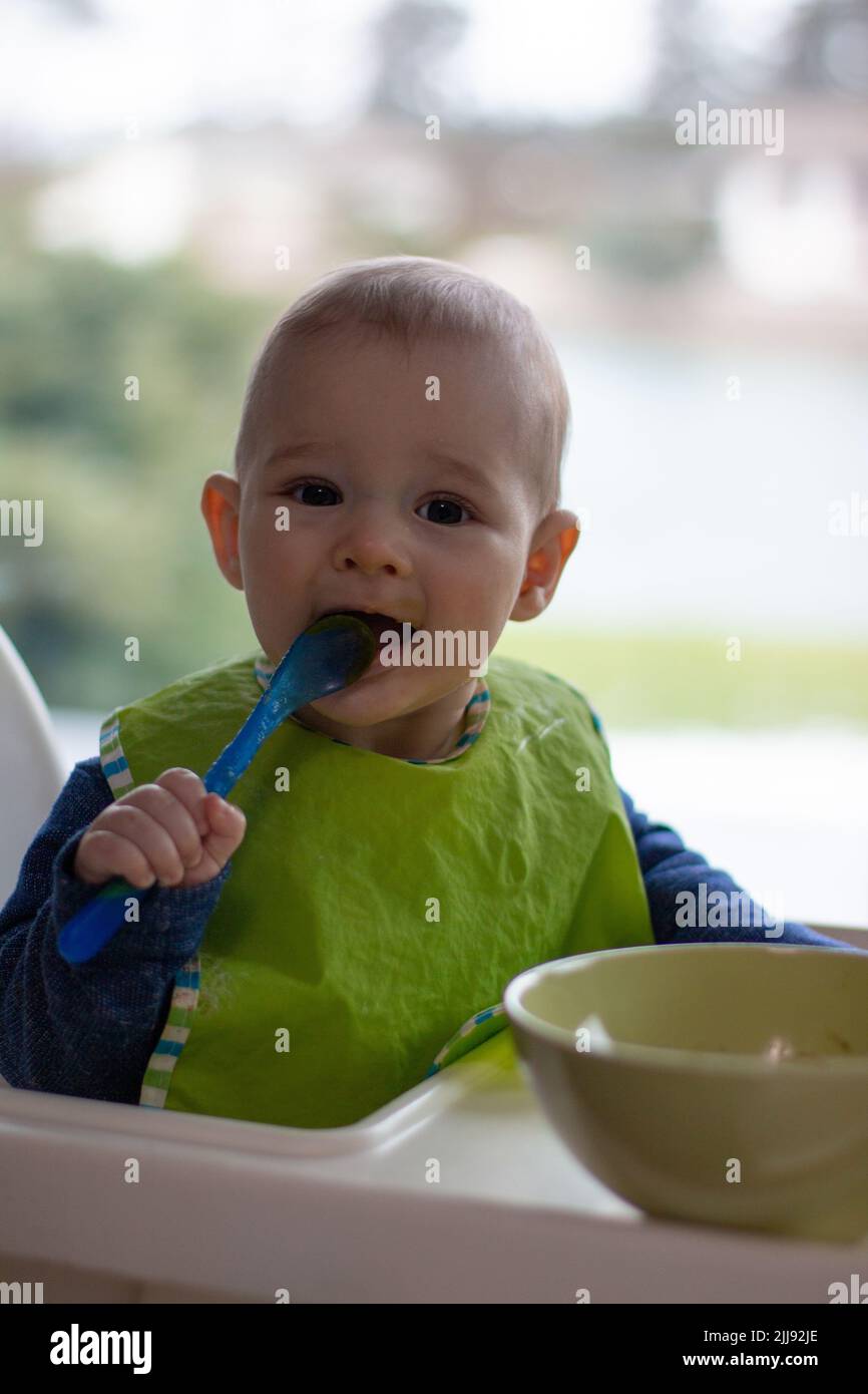Little toddler with a spoon on eating chair, happy baby boy learning to
