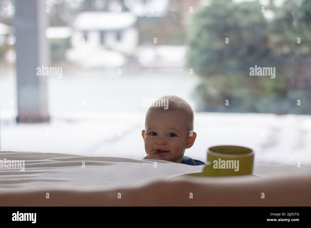 Curious, playful and mischievous child toddler with a finger in mount is watching to parents bed interested in a coffee cup, blonde boy in the bright Stock Photo