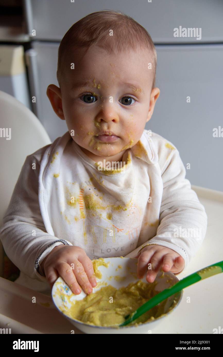 Messy and dirty baby eating food with hands and spoon on high chair
