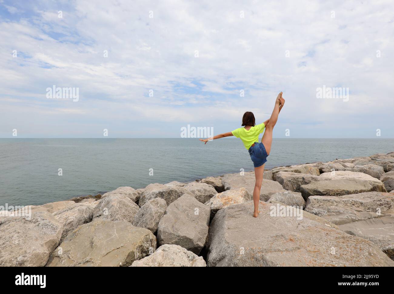 young girl does gymnastics exercises on the rocks by the sea in jeans ...