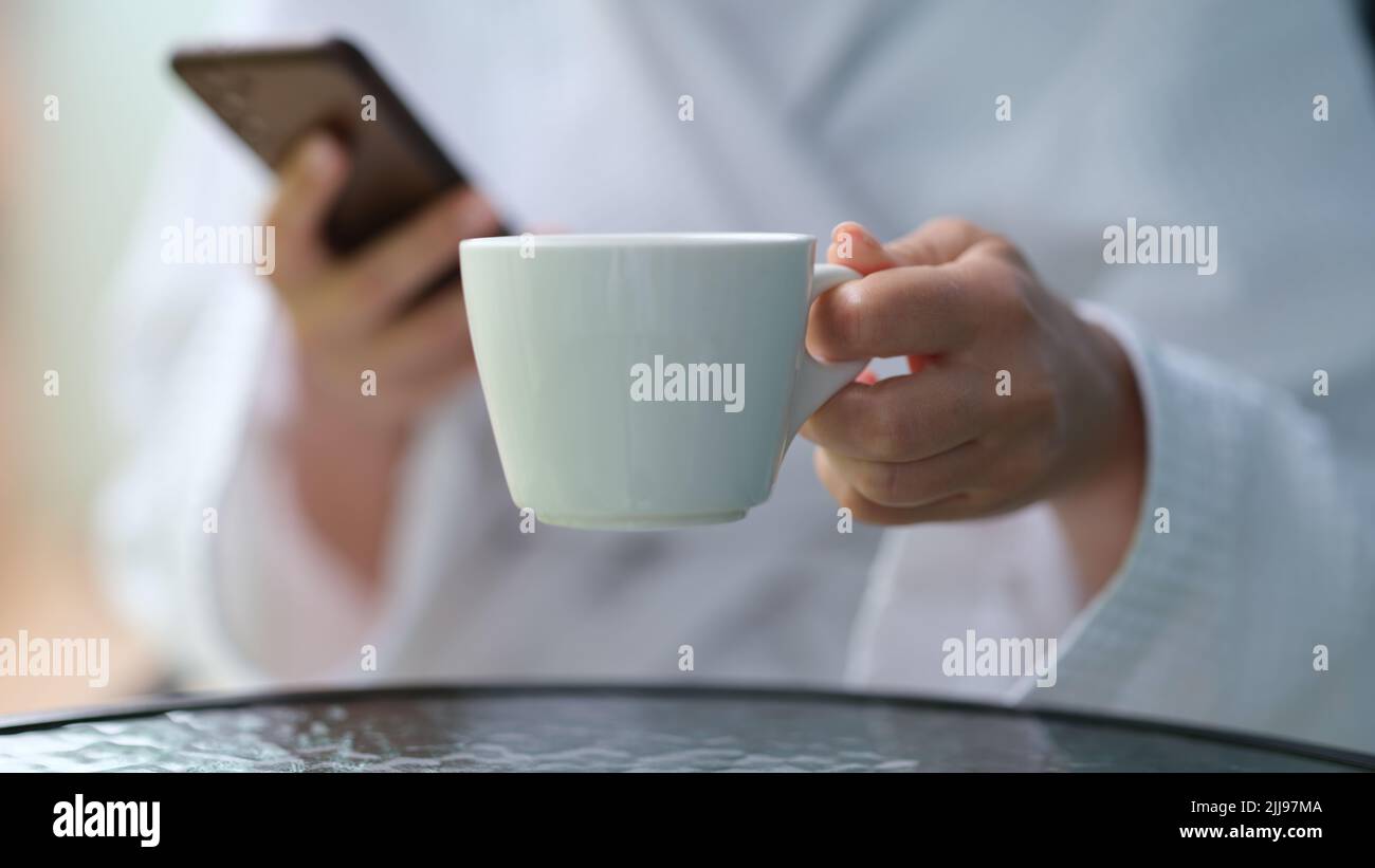 Cropped photo of a woman in a terry bathrobe holding mobile phone and cup Stock Photo