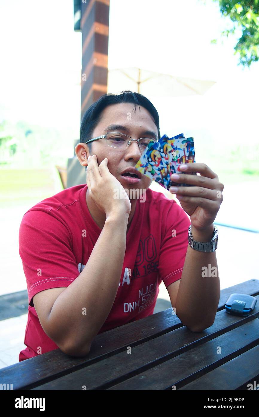 Portrait of young and handsome Asian man sitting and holding cards in poker game on wooden table. Serious expression. Stock Photo