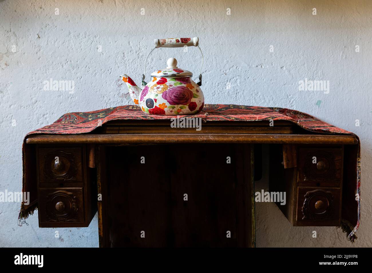 Old teapot decorated with hand-painted flowers, on an old table, with an embroidered mandel, on a white wall Stock Photo