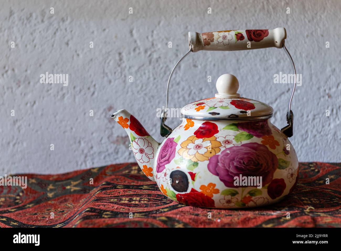 Old teapot decorated with hand-painted flowers, on an old table, with an embroidered mandel, on a white wall Stock Photo