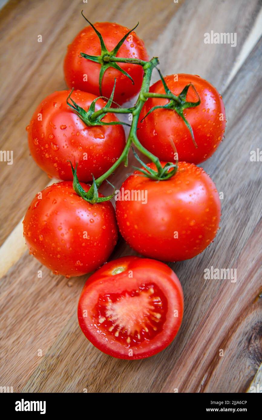 Red tomatoes on vine in kitchen - sliced and whole fresh tomato on wood cutting board - ripe Solanum lycopersicum bunch and cluster Stock Photo
