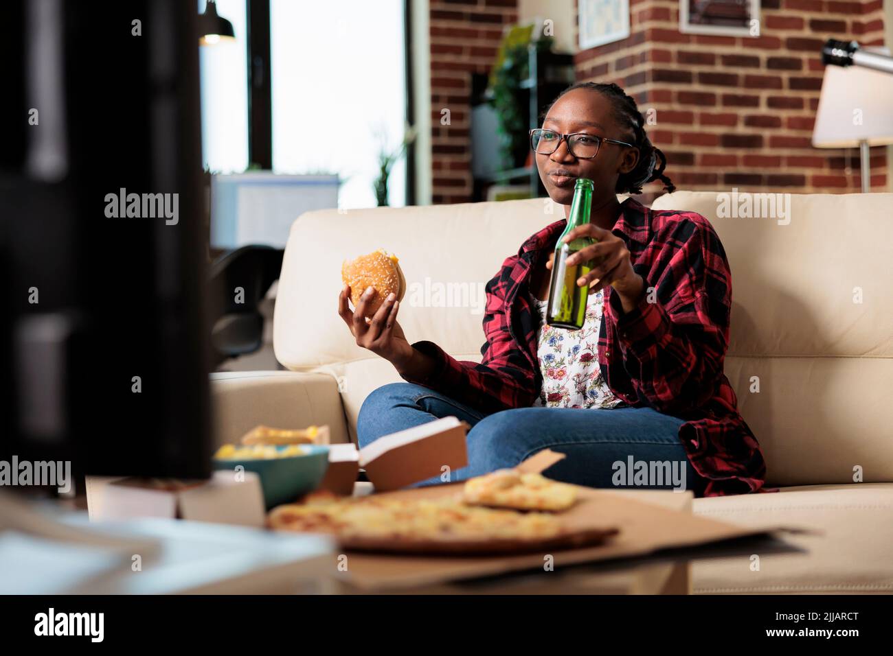 Smiling woman eating burger and drinking bottle of beer, enjoying leisure activity and fun movie on television. Having takeaway fast food delivery meal with beverage and snacks at home. Stock Photo