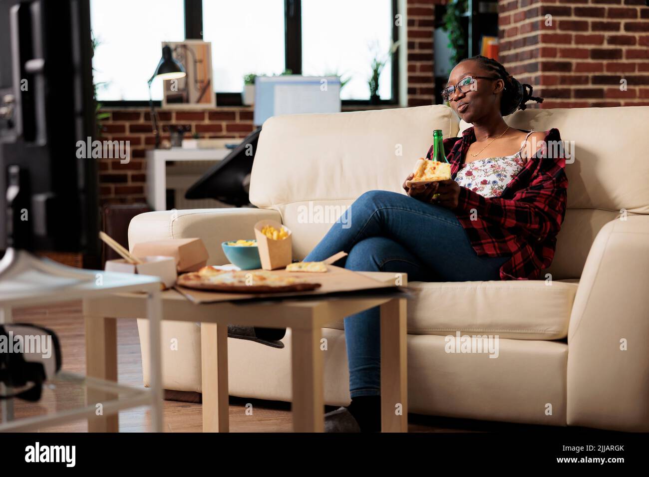 Young adult watching television with beer bottle and pizza in hand, enjoying takeaway delivery meal. Eating fast food takeout meal with alcoholic drink, looking at film on tv channel. Stock Photo