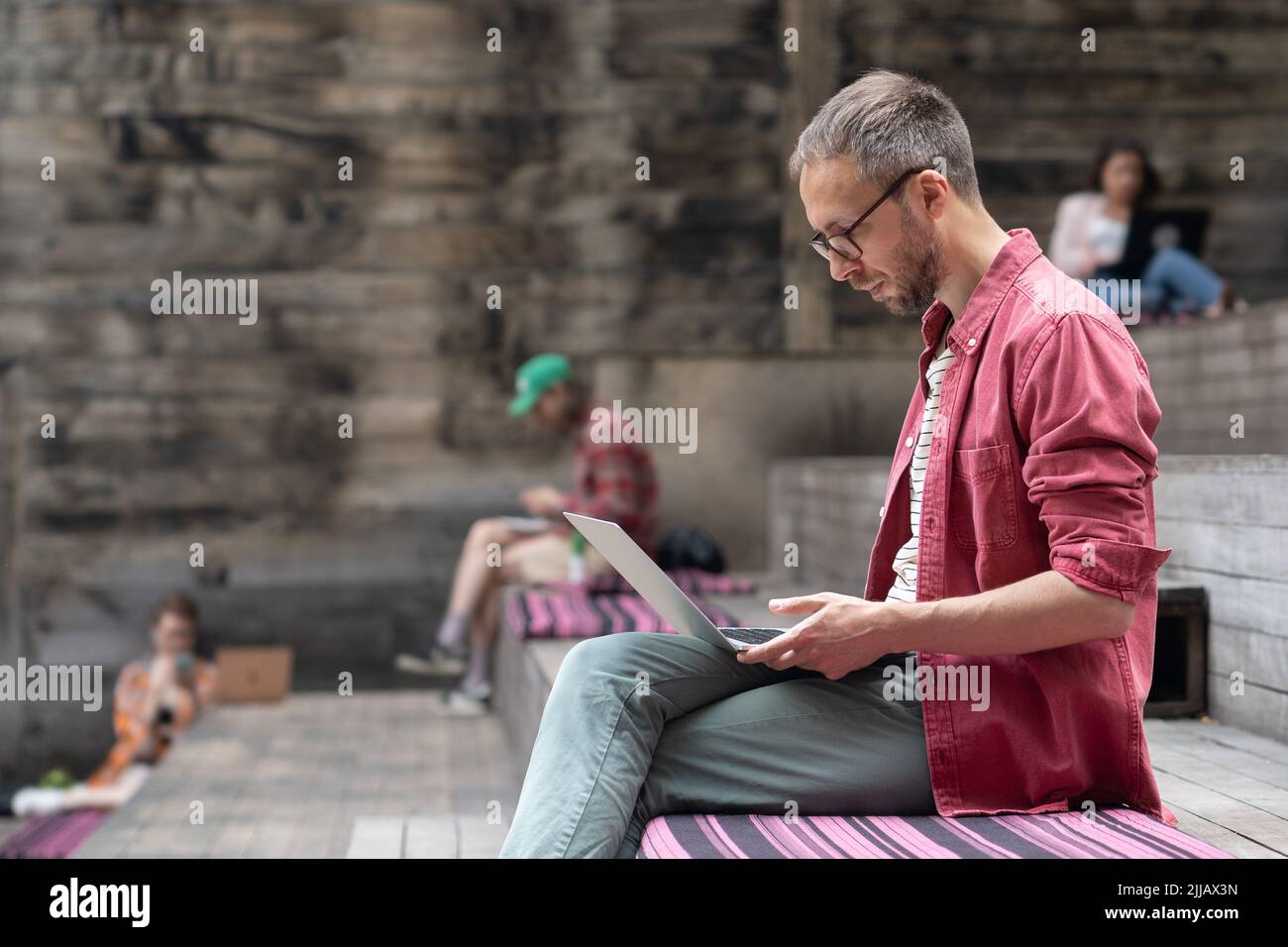 Young focused student guy with laptop sitting on wooden bench outside. Outdoor coworking space Stock Photo