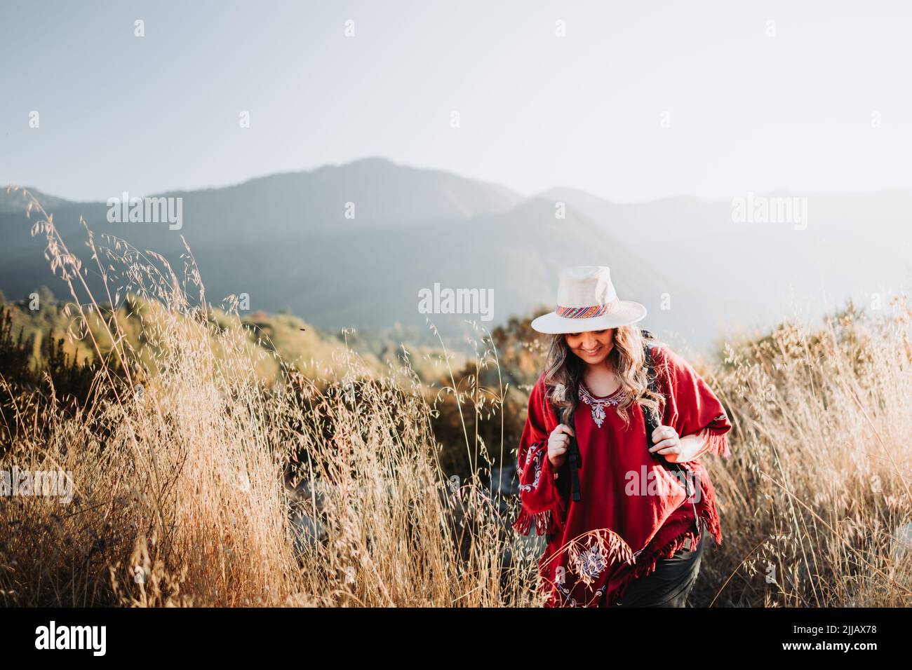 Latin backpacker woman wearing a traditional red poncho and a hat in a sunny landscape. Copy space Stock Photo