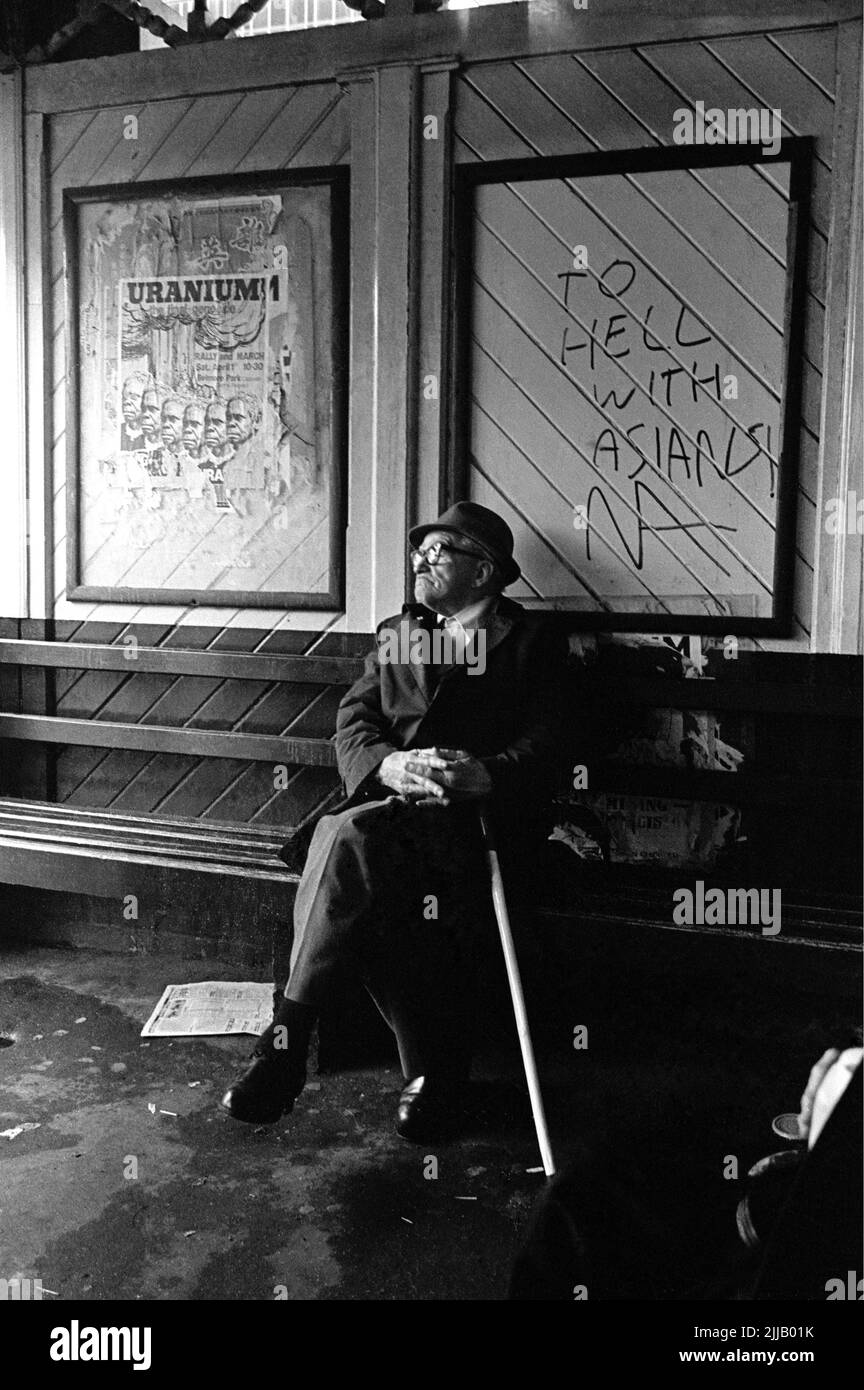 Old man with walking stick sitting in bus shelter with racist graffiti, saying 'To hell with Asians', and and anti-uranium mining poster in Sydney city CBD in 1977 Stock Photo