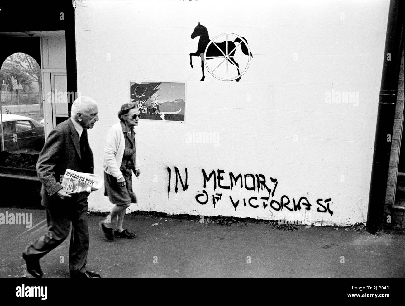 An elderly couple, residents of Sydney's Kings Cross walking in one of the side with activist green ban graffiti. Man carrying newspaper with headline, Arab Bloodbath. Vintage photo Stock Photo