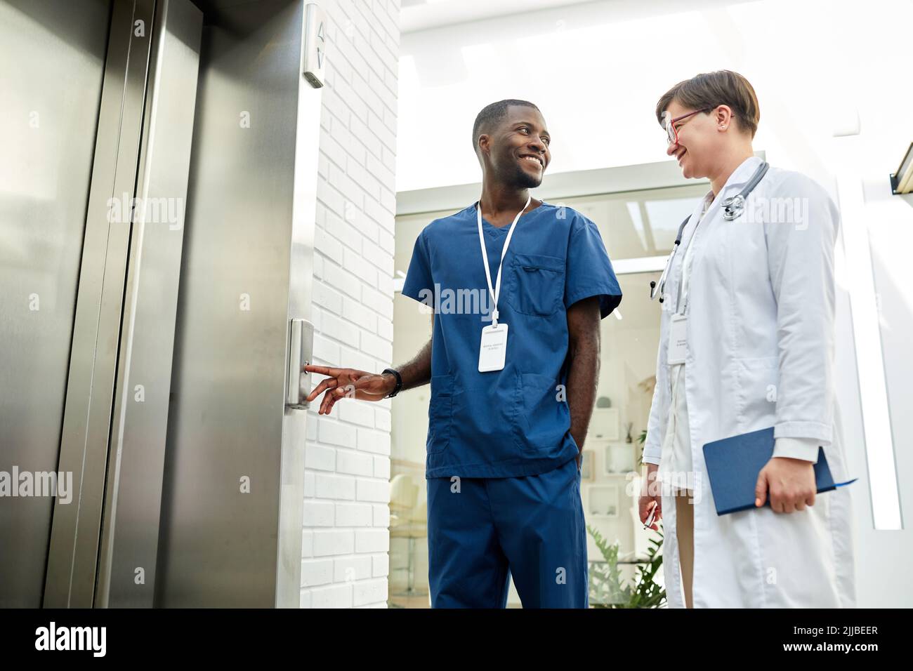 Low angle portrait of two medics waiting for elevator in modern hospital building and chatting cheerfully Stock Photo
