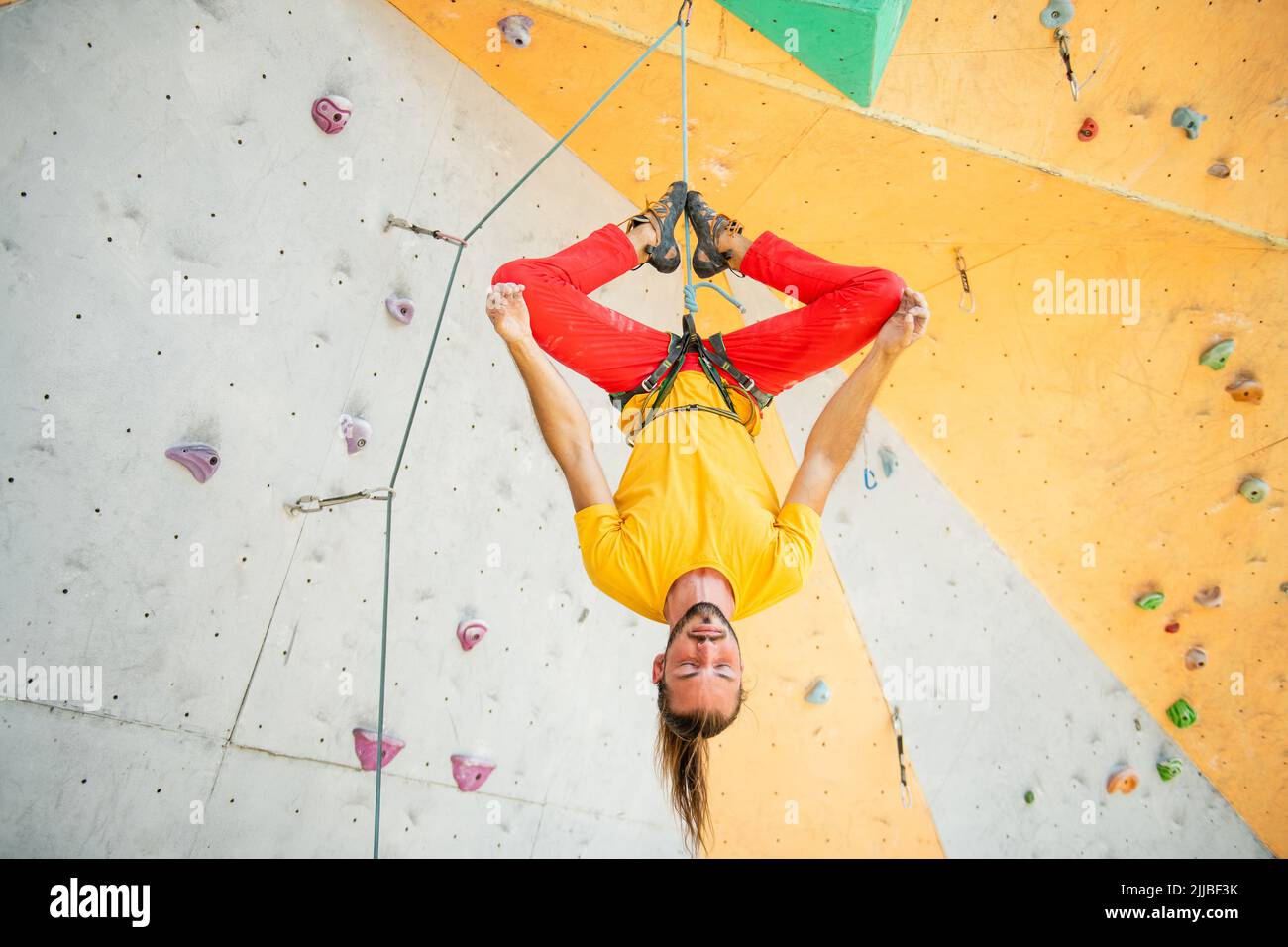 A man hangs upside down in a lotus position on a climbing wall. The man is wearing a yellow T-shirt and red pants. Stock Photo