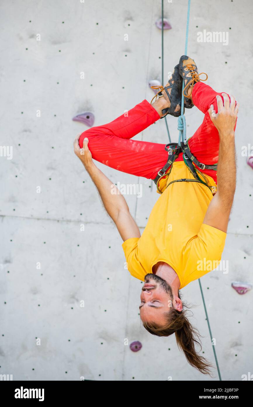 A man hangs upside down in a lotus position on a climbing wall. The man is wearing a yellow T-shirt and red pants. Stock Photo