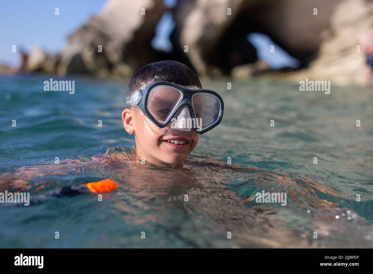 Happy young boy in goggles smiling in sea at rocky shore Stock Photo