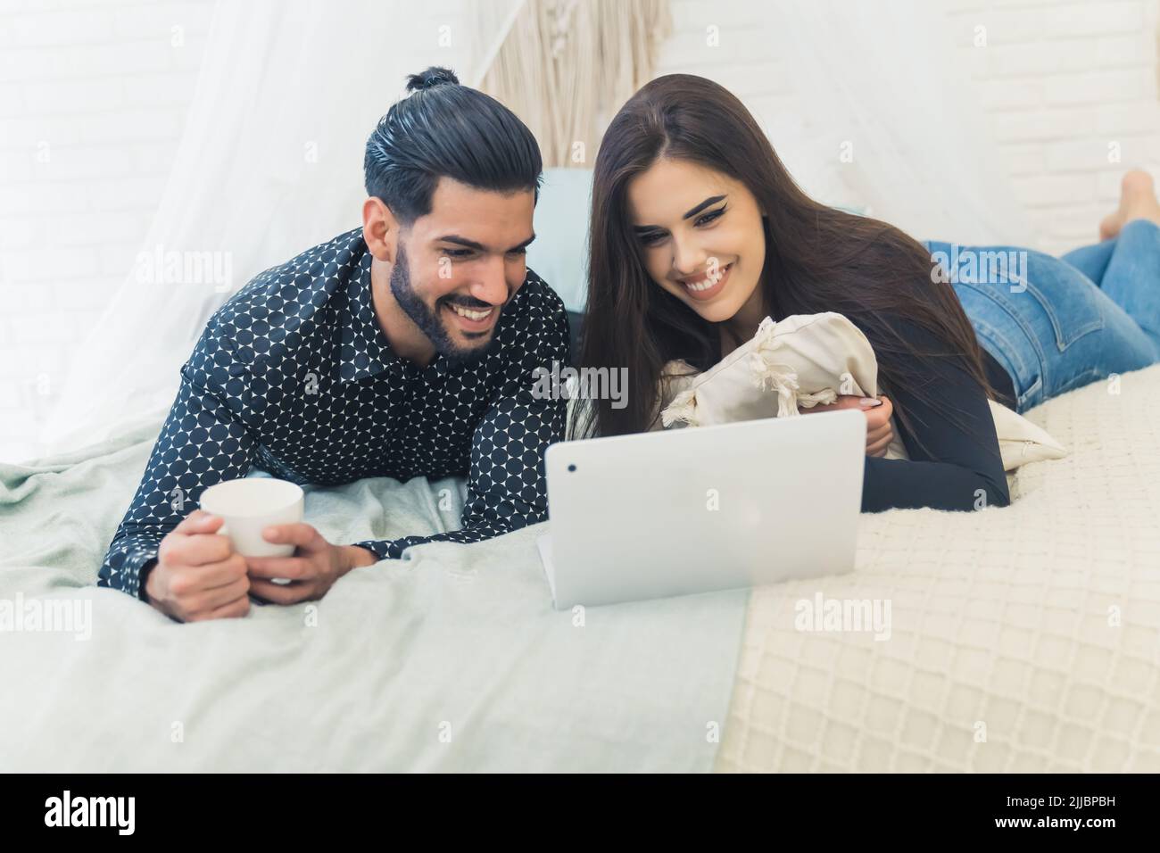 Cute couple watching film on laptop on bed with white sheets while man is holding a cup of coffee. Stock Photo