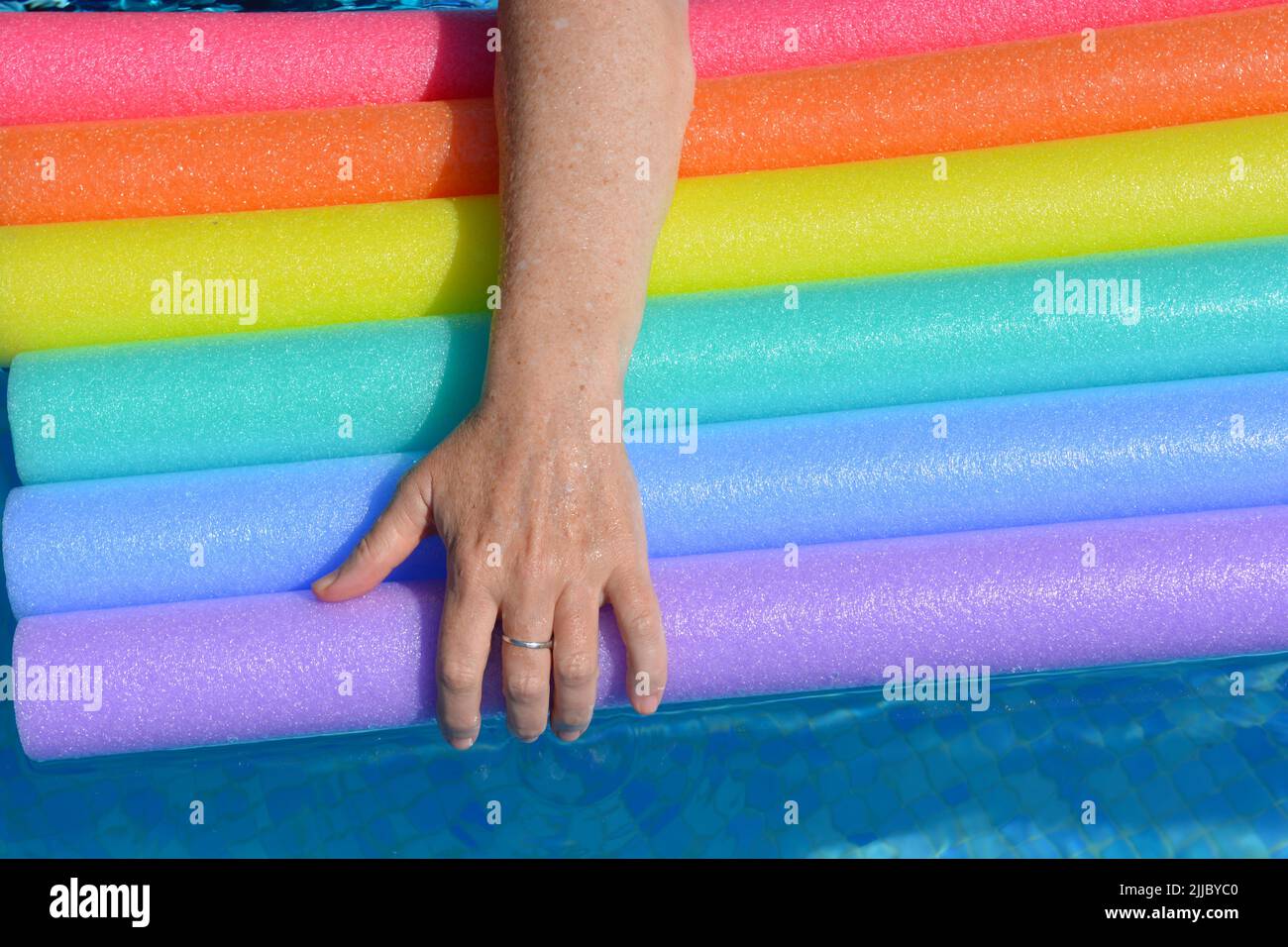 Woman's arm resting on rainbow coloured swim noodles in swimming pool Stock Photo