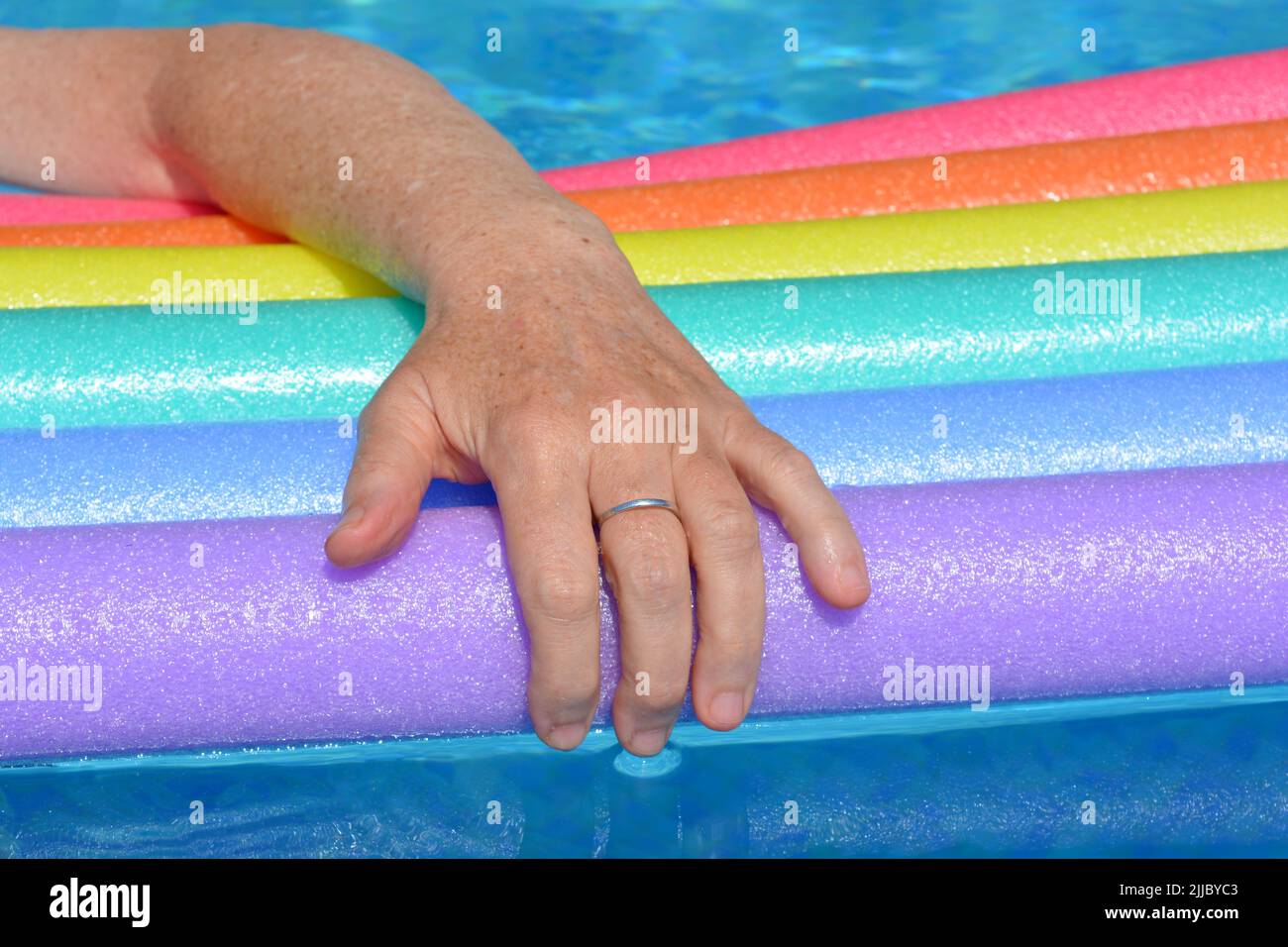 Woman's arm resting on rainbow coloured swim noodles floating in swimming pool Stock Photo
