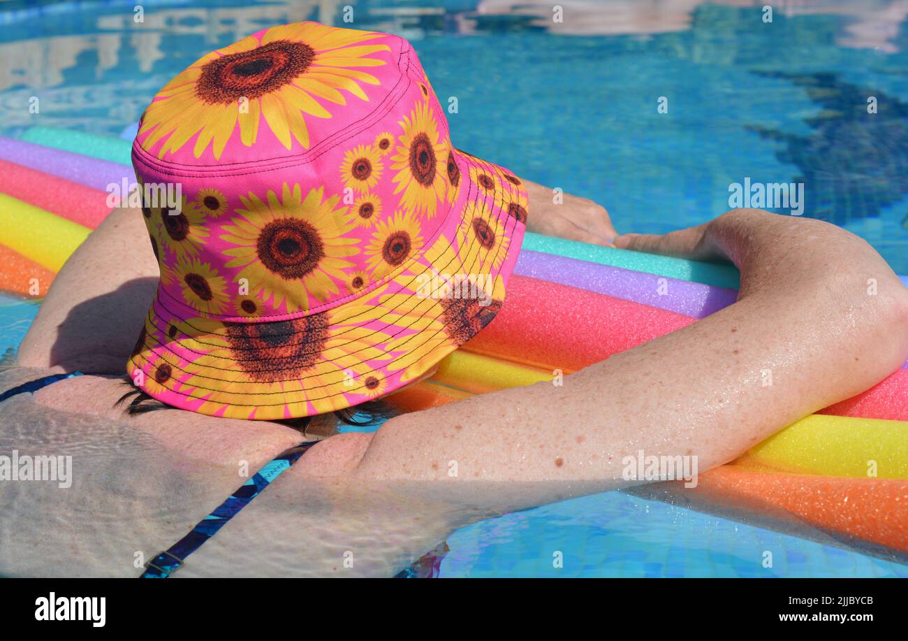 Woman wearing sun hat floating on swim noodles in swimming pool, from behind Stock Photo