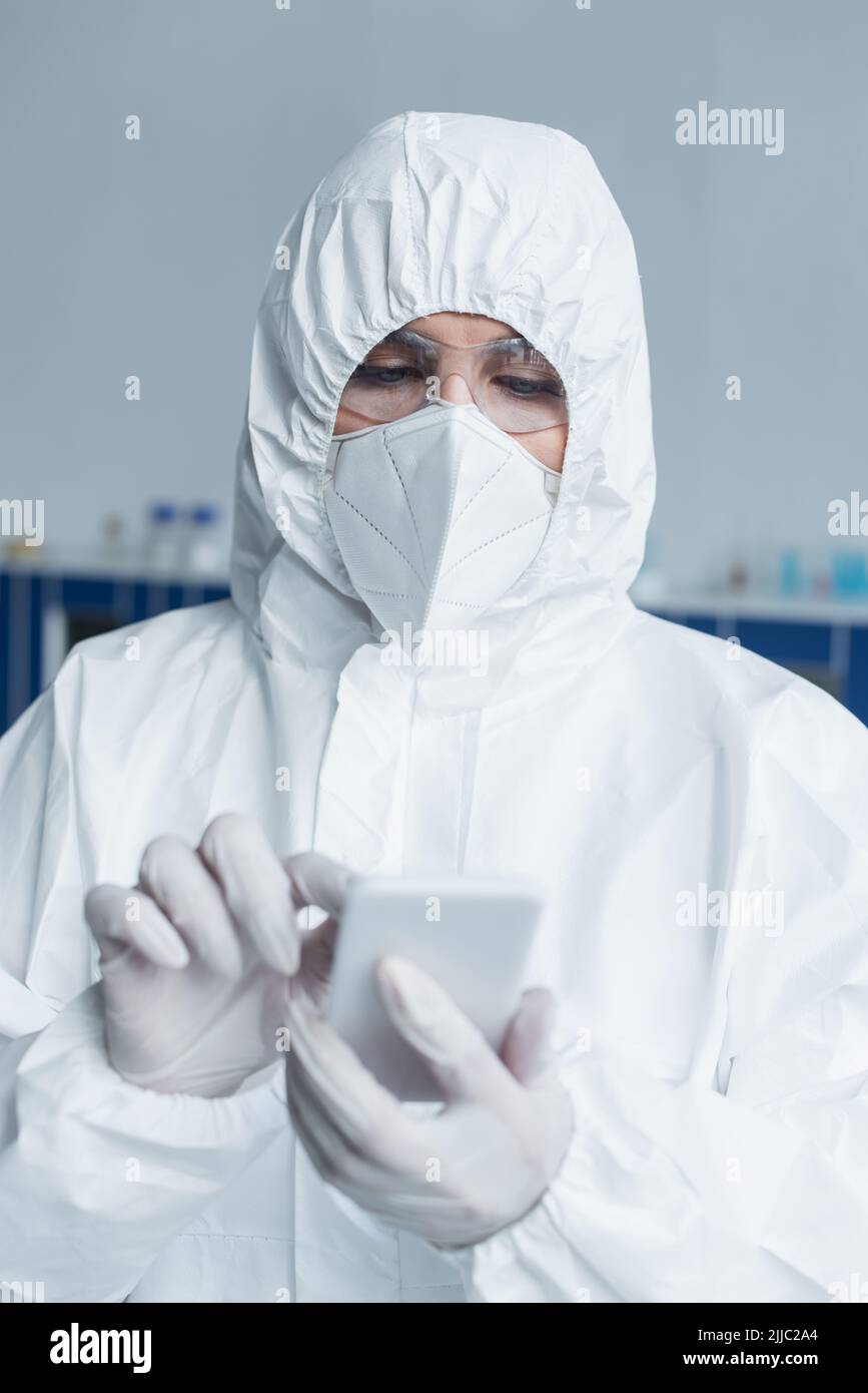 Scientist in hazmat suit and protective mask using blurred smartphone in lab Stock Photo